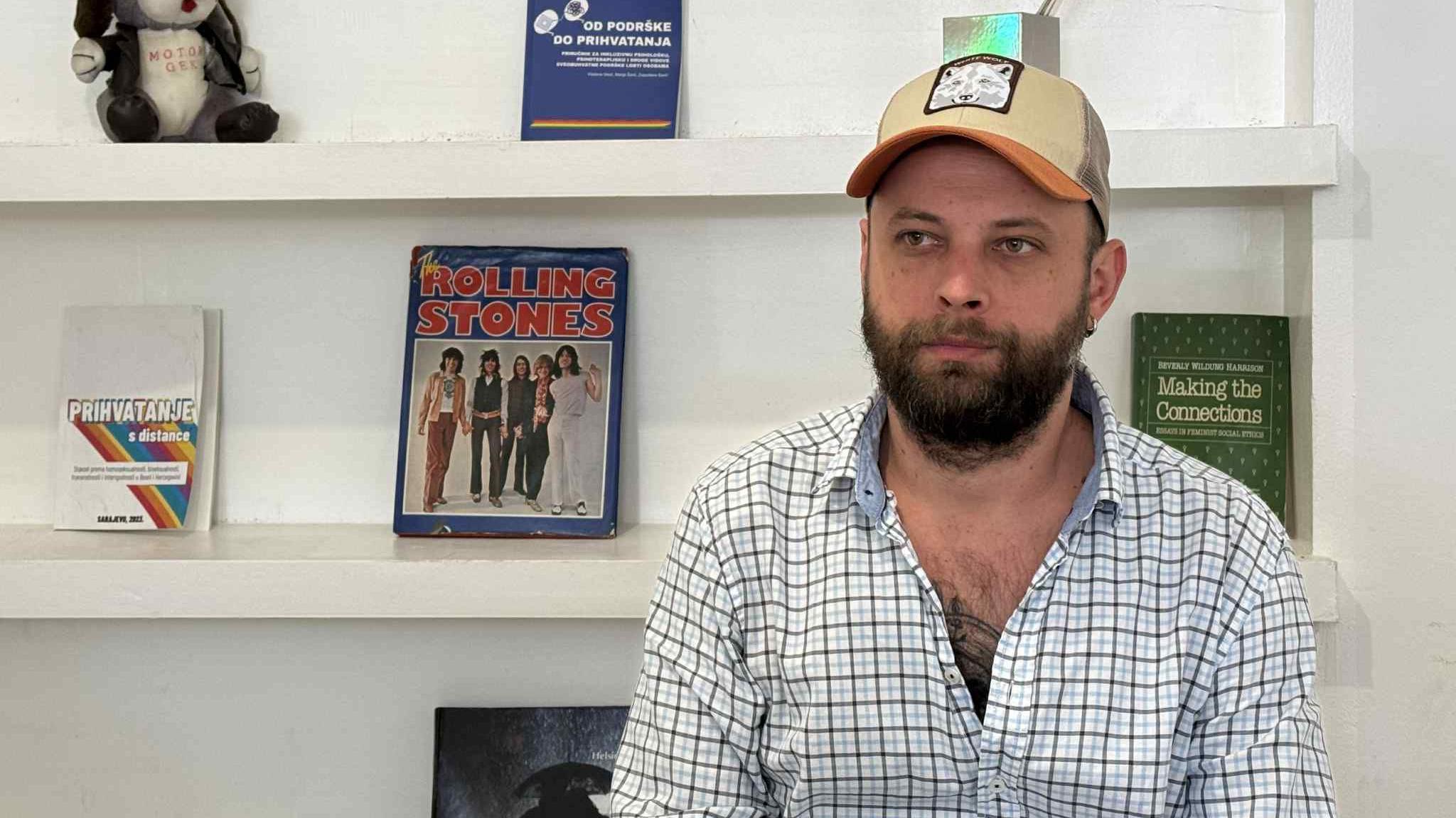 Muškarac sa bradom i kačketom na glavi, sedi u kariranoj beloj košulji na plave i crne pruge, iza njega su police sa časopisima i knjigama