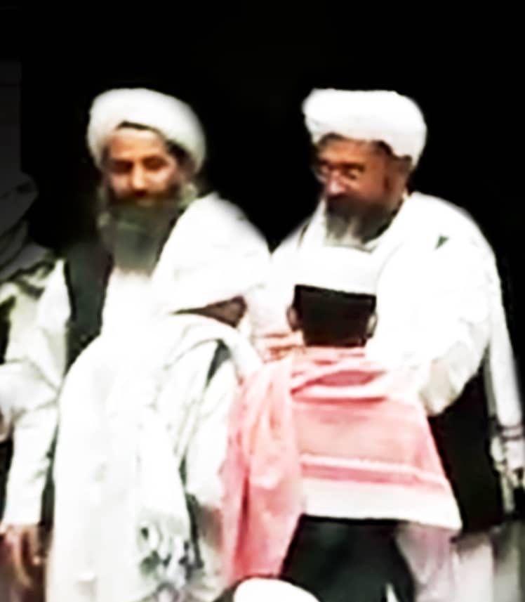 A grainy image showing two men dressed in white with turbans. The man on the left has a large beard which is greying. The man on the right wears glasses, with a slightly shorter beard. In front of them are what appears to be two boys. File photo