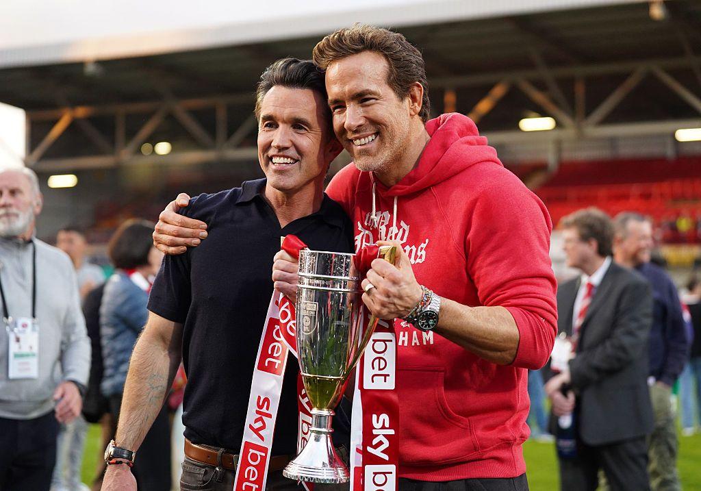 Rob McElhenney e Ryan Reynolds sorrindo e posando para foto segurando troféu; eles estão no gramado