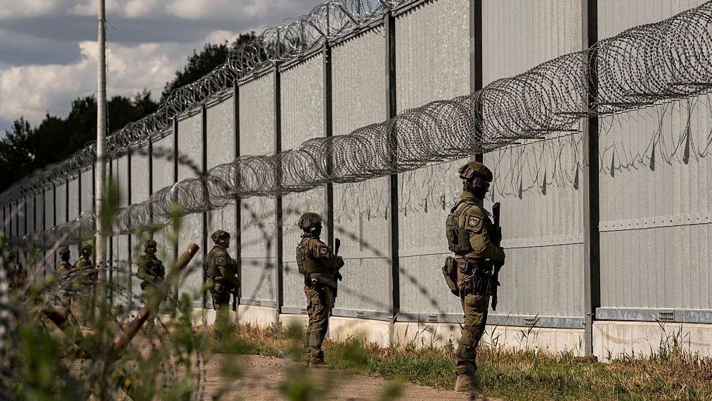Soldados fronterizos armados, montan guardia frente a una larga y alta valla metálica con concertinas de alambres de púas