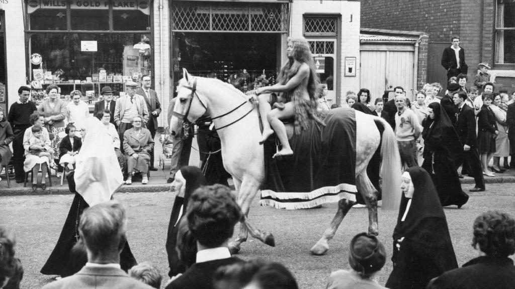 Una mujer desnuda y con el cabello muy largo pasea en un caballo en un desfile por la calle de Reino Unido. Hay personas mirando y varias monjas acompañan el desfile. La foto es en blanco y negro.