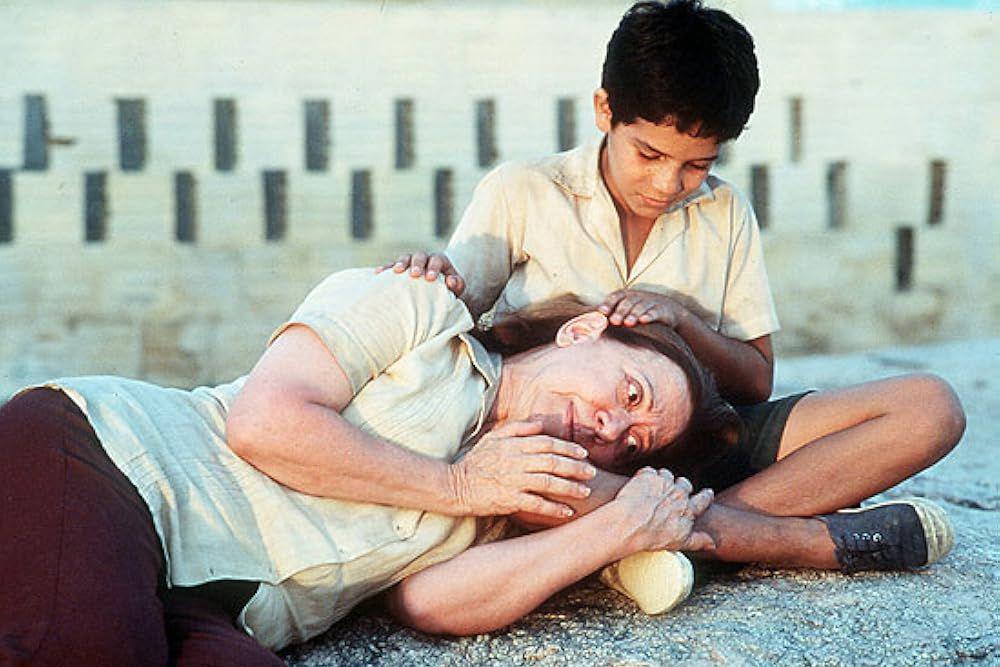 A atriz Fernanda Montenegro aparece deitada no ch&atilde;o, com express&atilde;o abatida e olhar distante, enquanto o ator Vin&iacute;cius de Oliveira est&aacute; sentado atr&aacute;s dela, apoiando a m&atilde;o sobre sua cabe&ccedil;a. Ela veste uma blusa clara, enquanto ele usa camisa bege e shorts escuros. O cen&aacute;rio ao fundo &eacute; &aacute;rido, com postes fincados na paisagem seca.