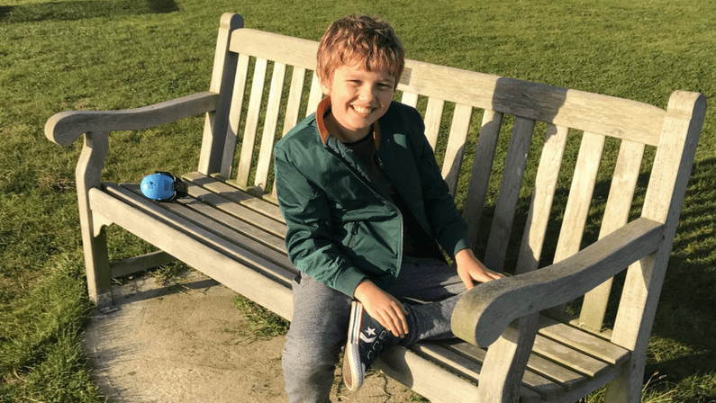 Jude sorri para a câmera. Ele está sentado em um banco, em um parque, e usa uma jaqueta verde.