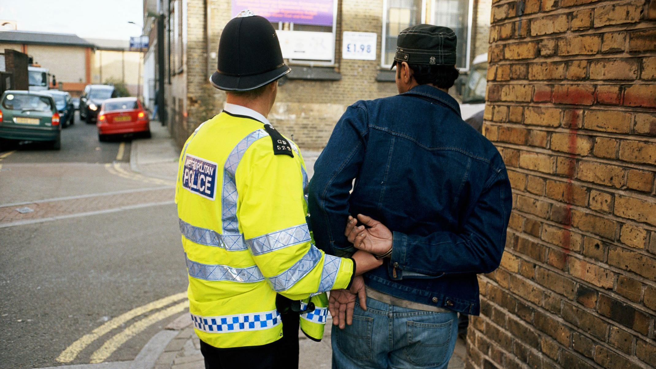 Policial de costas ao lado de homem algemado, tamb&eacute;m de costas; eles est&atilde;o em rua