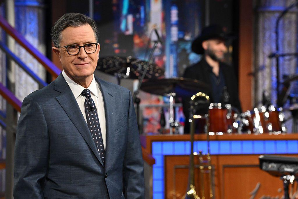 Stephen Colbert de pie en el set; detrás aparece una batería y un músico desenfocado.