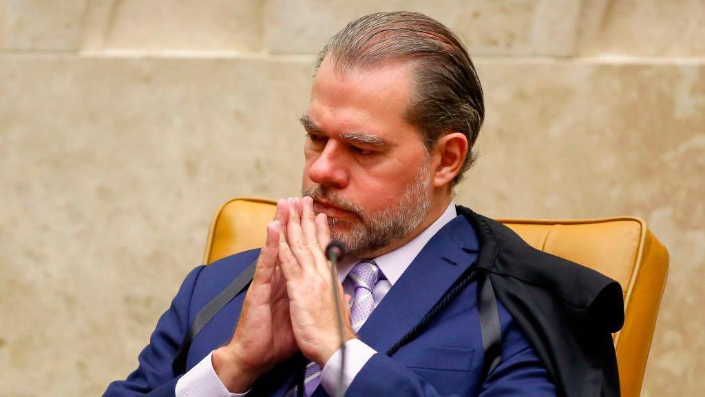 Dias Toffoli, um homem de meia-idade, com cabelos penteados para tr&aacute;s e barba grisalha bem aparada, est&aacute; sentado em uma cadeira de couro em um ambiente que parece institucional. Ele veste terno azul com gravata clara e tem um casaco ou toga apoiado sobre os ombros. Com as m&atilde;os unidas diante do rosto, em gesto de concentra&ccedil;&atilde;o, mant&eacute;m os olhos semicerrados.