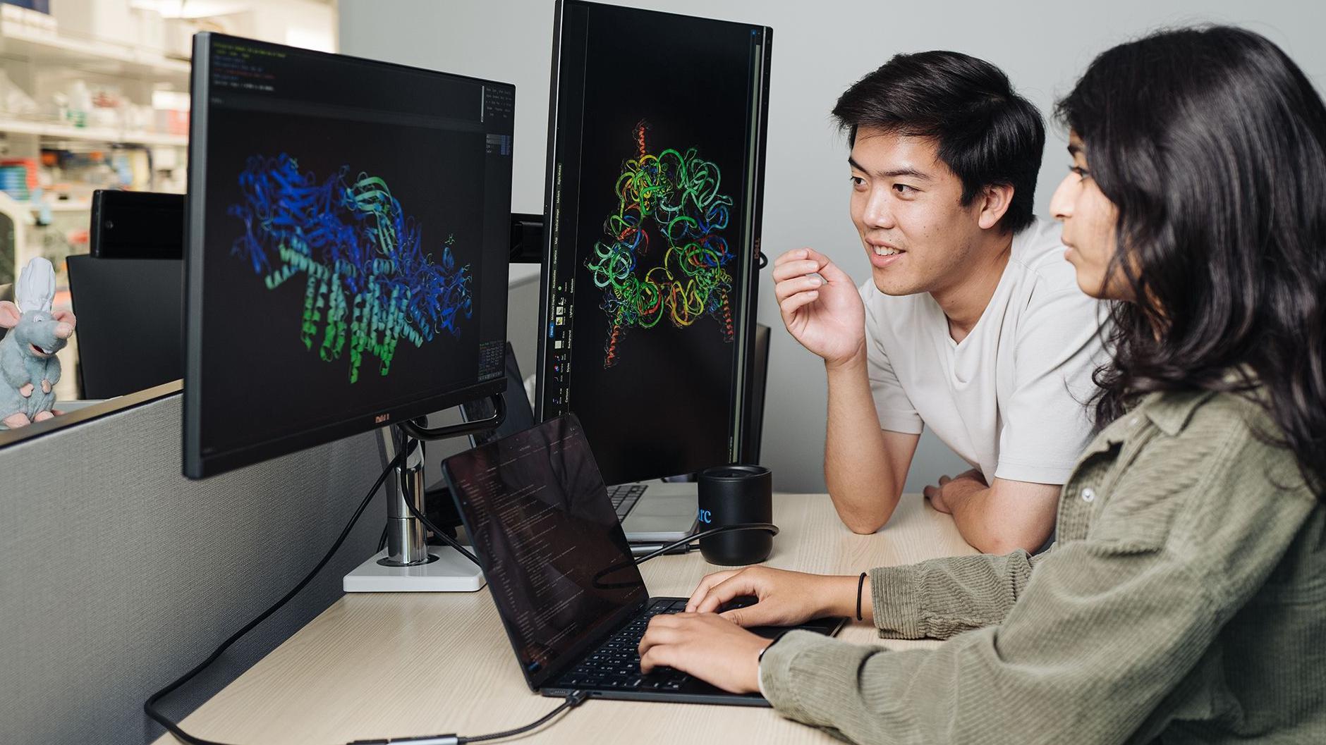 Dos personas trabajan juntas en un escritorio, aparentemente en un laboratorio o centro de investigación. Grandes monitores muestran coloridos modelos tridimensionales de estructuras moleculares o proteicas sobre fondos oscuros, mientras que un portátil sobre el escritorio muestra líneas de código o datos. Una persona está sentada usando el portátil y la otra se inclina hacia las pantallas, ambas concentradas en las visualizaciones científicas. Al fondo se aprecian estanterías y equipos de laboratorio, lo que sugiere un entorno de investigación biomédica, biotecnológica o computacional. La escena enfatiza el uso de herramientas de análisis y visualización informáticas para estudiar estructuras biológicas complejas.