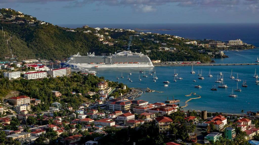 Panorama atual do porto de Saint Thomas, com pequenas embarca&ccedil;&otilde;es e um grande navio de cruzeiro em um mar azul e um c&eacute;u igualmente azul.