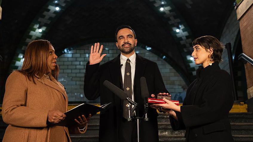 Zoran Mamdani, de barba, corbata y abrigo oscuro, levanta la mano derecha al ser juramentado por la fiscal general de Nueva York Letitia James (a la izquierda) que sostiene un documento. Rama Duwaji (a la derecha), una mujer con abrigo oscuro lo mira atentamente y sostiene un Corán donde Mamdami apoya su mano izquierda.