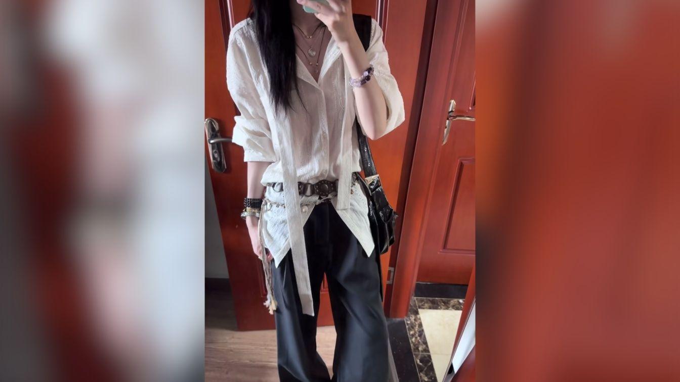 Mujer de pie haciéndose un selfie frente a un espejo con una pared roja en el fondo, vestida con pantalones negros y camisa blanca con flecos largos