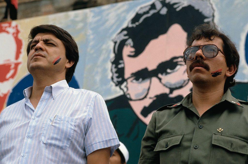 Una foto de 1984 que muestra al lado izquierdo a Sergio Ramírez vestido con una camisa de rayas. A su derecha está Daniel Ortega, vestido de uniforme militar. Ambos miran al horizonte y tienen una mejilla pintada con una franja roja y negra.