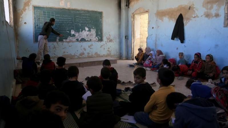 أطفال من البنين والبنات وأماهم معلم يشرح على سبورة في مدرسة في سوريا