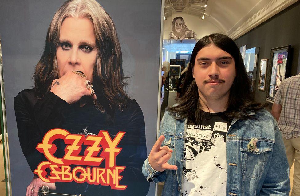 Um fã fazendo o sinal de rock com a mão, em pé ao lado de uma grande foto de Ozzy Osbourne