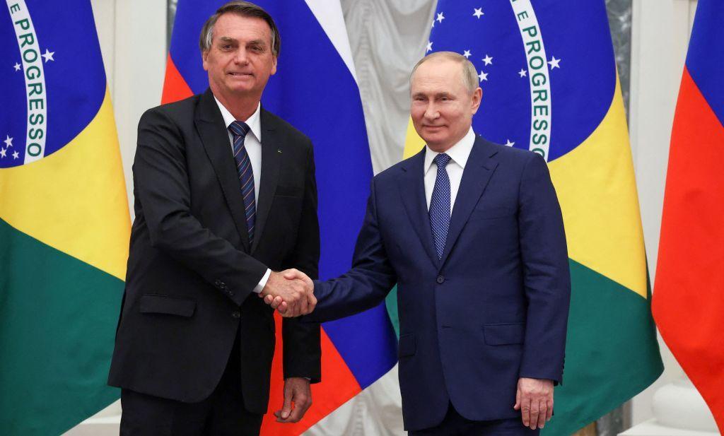 Bolsonaro e Putin sorrindo e apertando as mãos, em frente a bandeiras do Brasil e Rússia