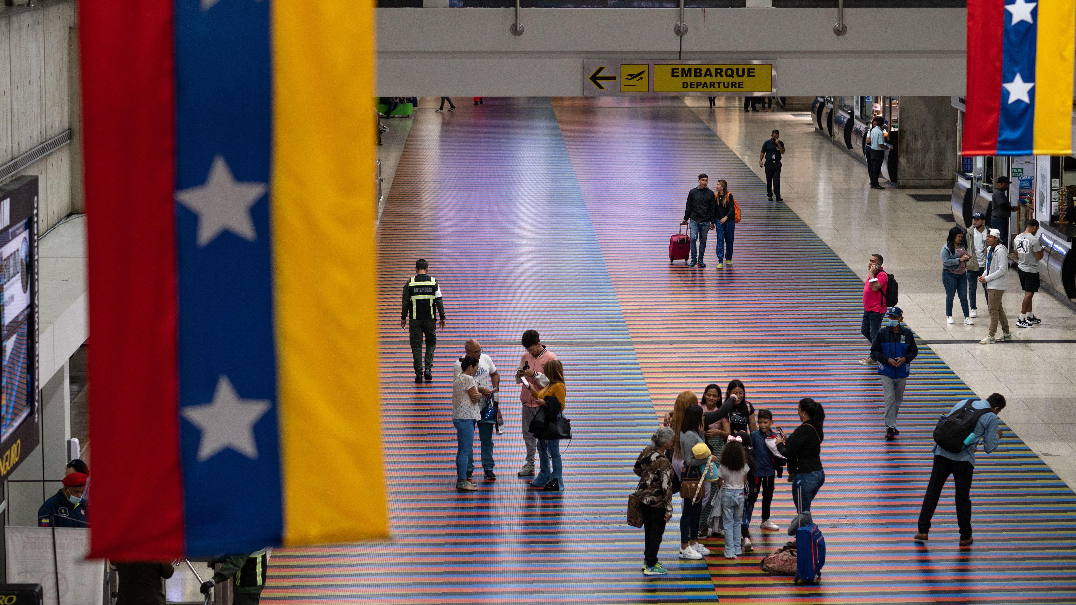 Vista interior del Aeropuerto Internacional de Maiquetía. A los lados hay dos pendones que cuelgan el techo con la bandera de Venezuela y en el centro se ve un pequeño grupo de personas de pie sobre un piso de muchos colores.