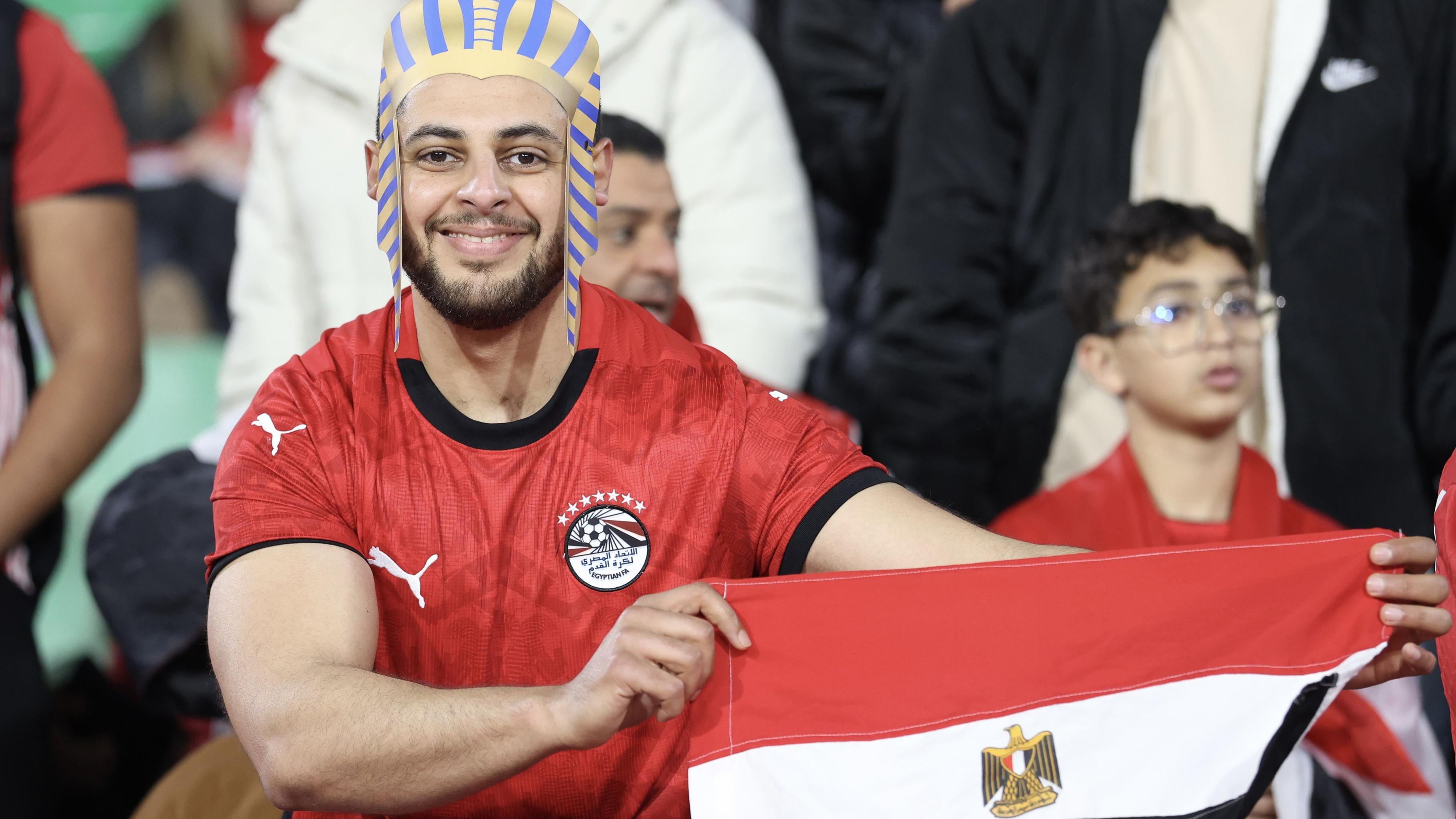 يظهر مشجع مصري مبتسم، يرتدي قميص المنتخب الوطني لكرة القدم باللون الأحمر وغطاء رأس على الطراز الفرعوني، وهو يرفع العلم المصري. ويمكن رؤية مشجعين آخرين غير واضحين في الخلفية.