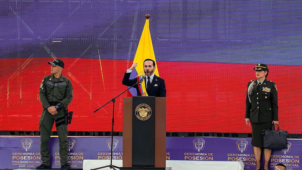De la Espriella faz seu discurso de posse em um palco em frente a uma tela exibindo a bandeira colombiana e acompanhado por dois representantes da for&ccedil;a p&uacute;blica, um homem e uma mulher.