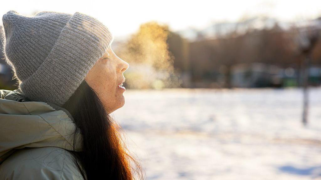 Mulher sentada ao ar livre, de lado, com os olhos fechados e roupas de frio