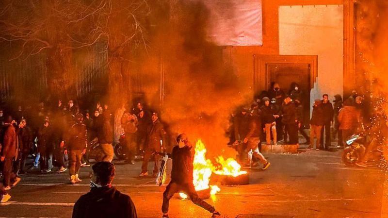 Fogo em uma rua de uma cidade iraniana e manifestantes com o rosto coberto