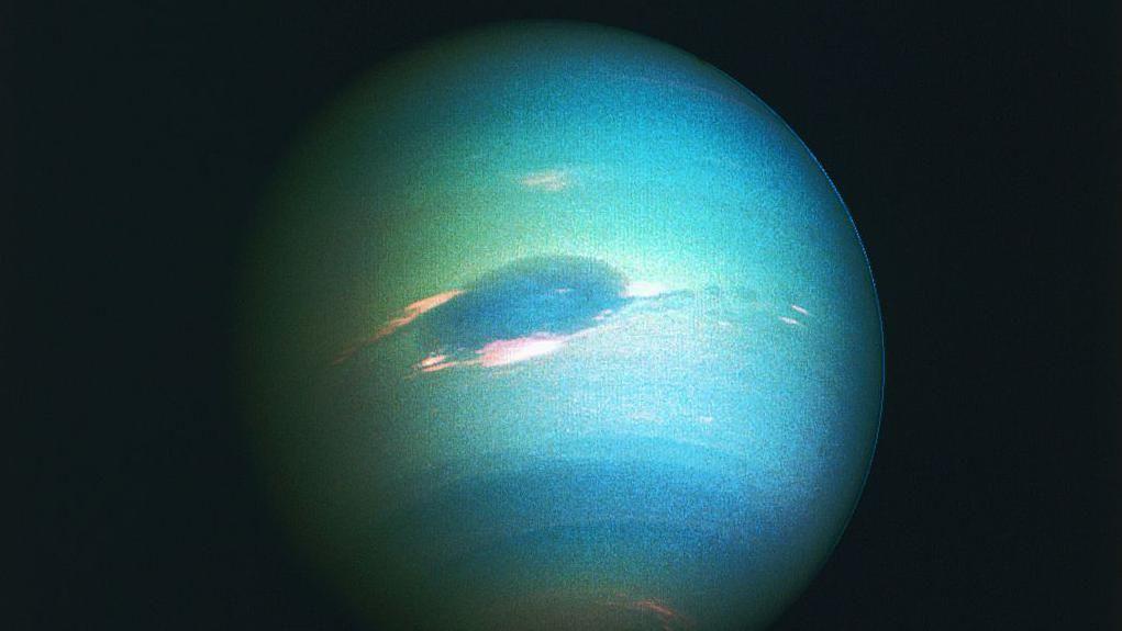 Neptuno se observa iluminado como una esfera casi completa, con diferentes tonalidades de bandas azul verdosas en su superficie y una gran área ovalada de color azul intenso en el centro.