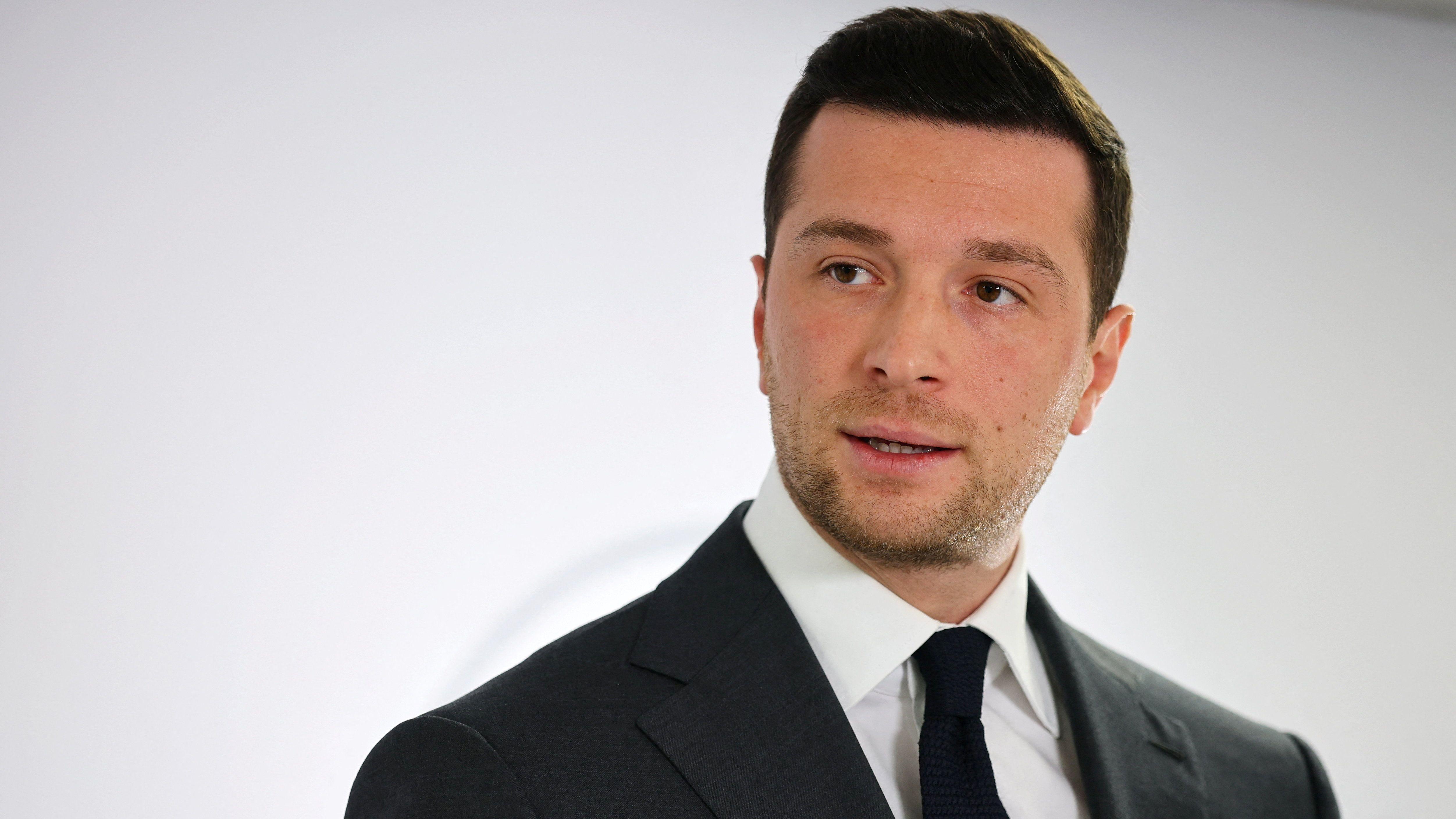 Jordan Bardella, con la cabeza ligeramente ladeada, viste traje y corbata oscuros y camisa blanca.