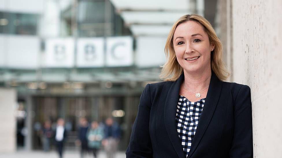 Zoe Kleinman con una blusa a cuadros blanca y negra y una chaqueta negra frente a la sede de la BBC en el centro de Londres