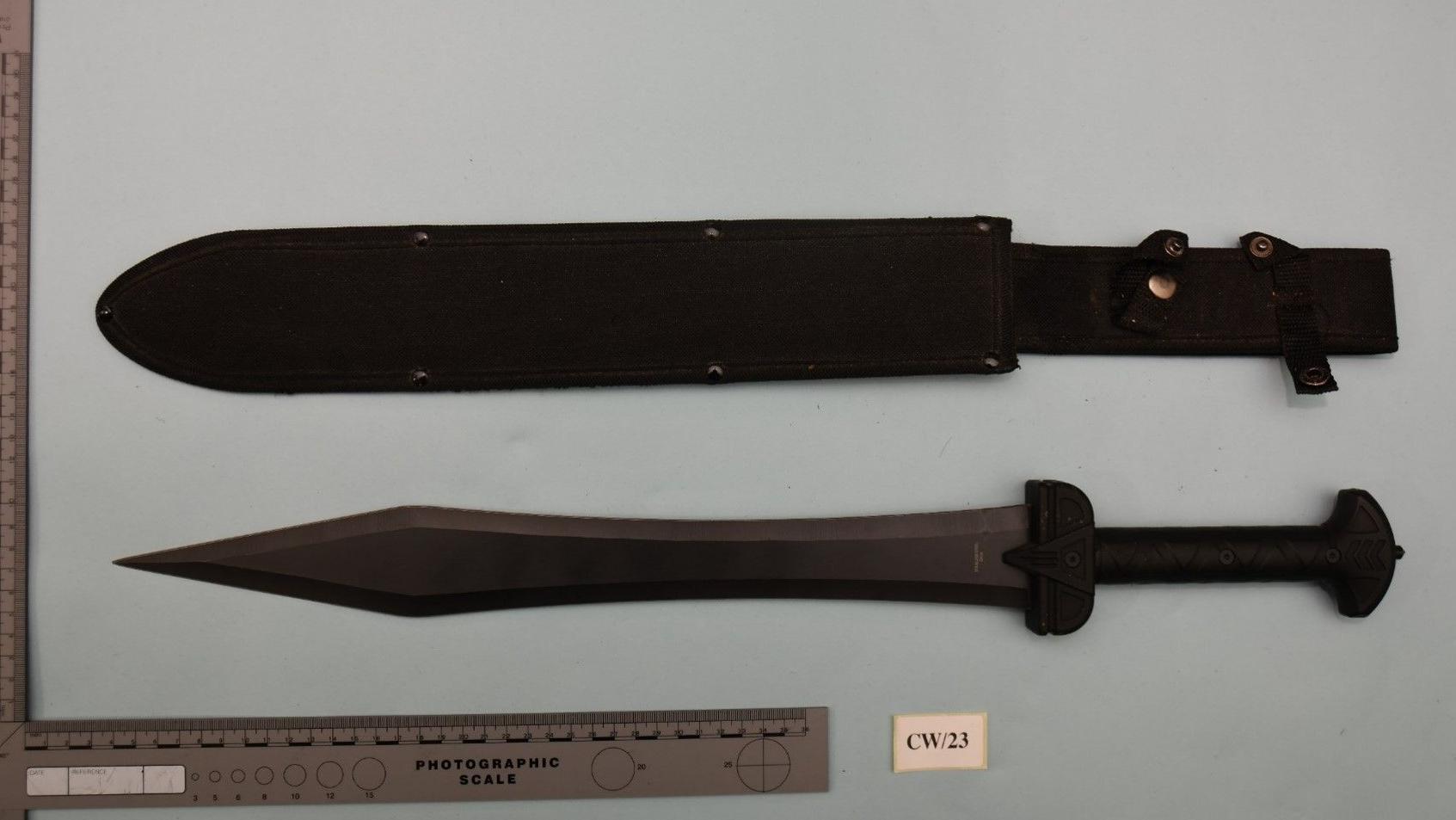 Uma espada colocada sobre um lençol de cor clara ao lado de um estojo usado para guardar a arma. Há uma régua ao lado da espada