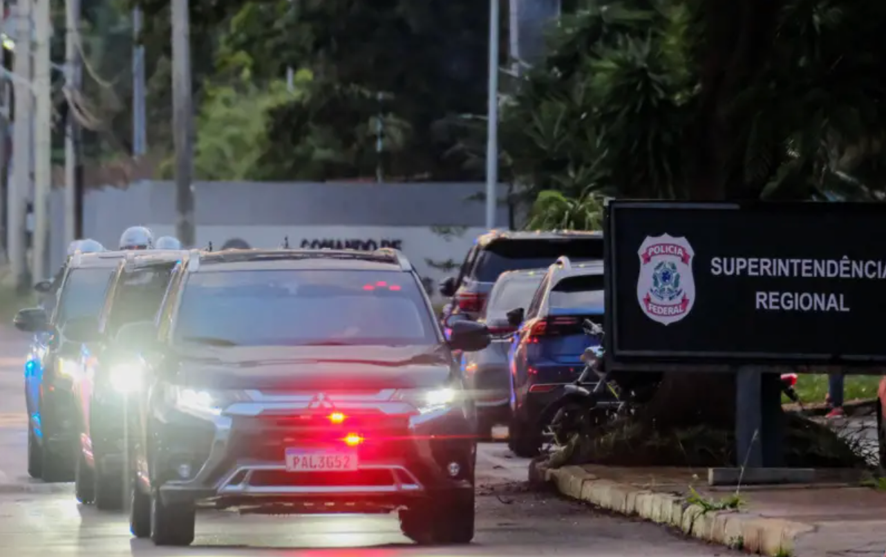 Foto mostra viaturas com farois e luzes de emerg&ecirc;ncia em frente a uma placa da Pol&iacute;cia Federal