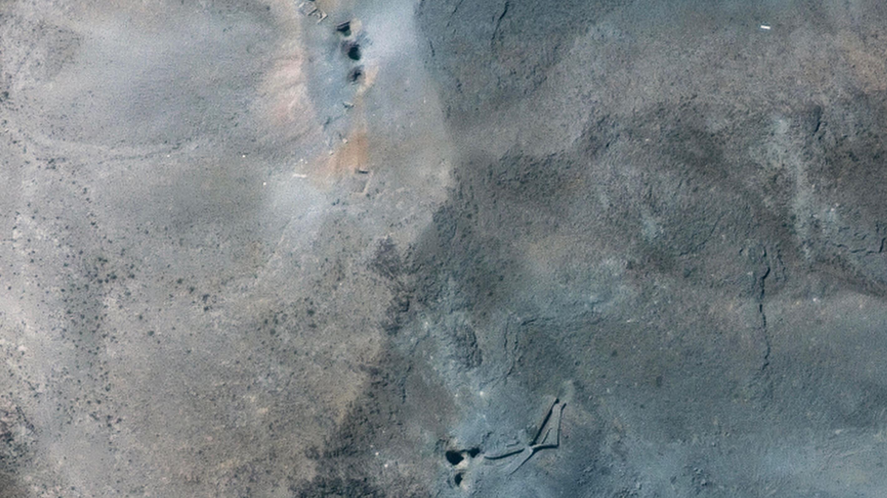 A handout satellite image made available by Maxar Technologies shows a close-up view of holes and craters on a ridge at the Fordo underground uranium enrichment site following US airstrikes. Three circular craters are in a straight line at the top of the image and three forming a triangle are at the bottom. The surrounding ground is grey and brown.