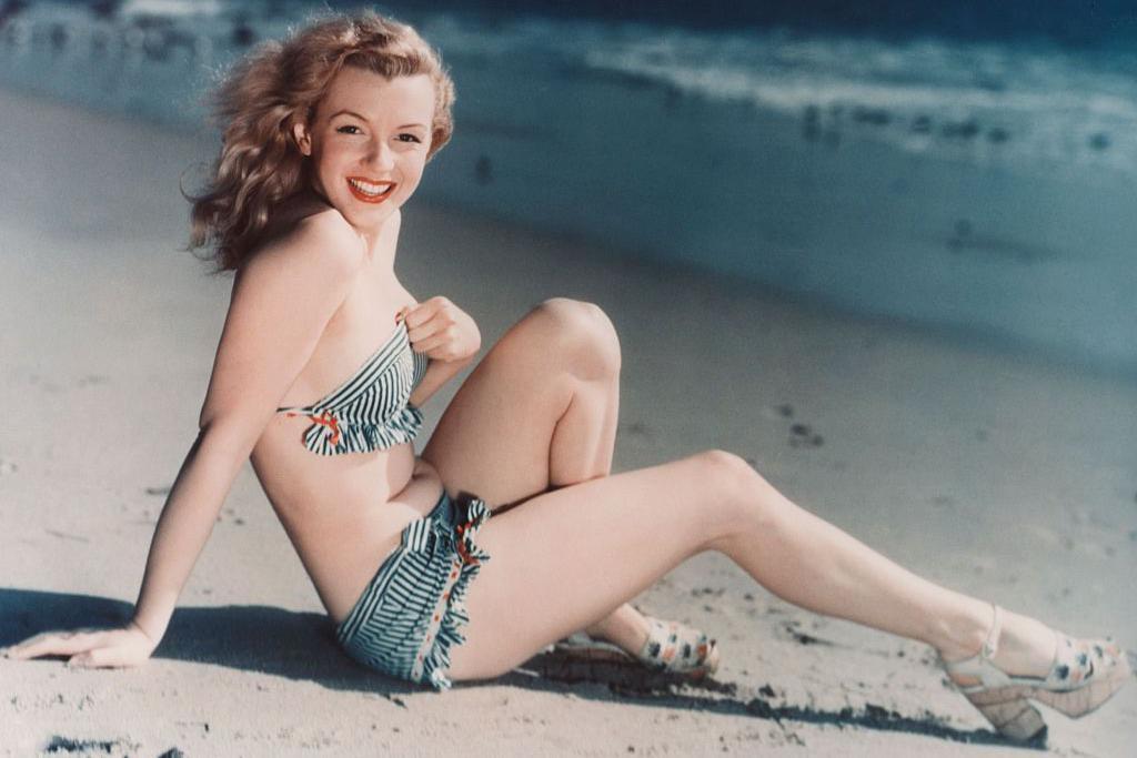 A jovem Marilyn Monroe, sentada na praia e sorrindo, com um biqu&iacute;ni verde estampado e sand&aacute;lias