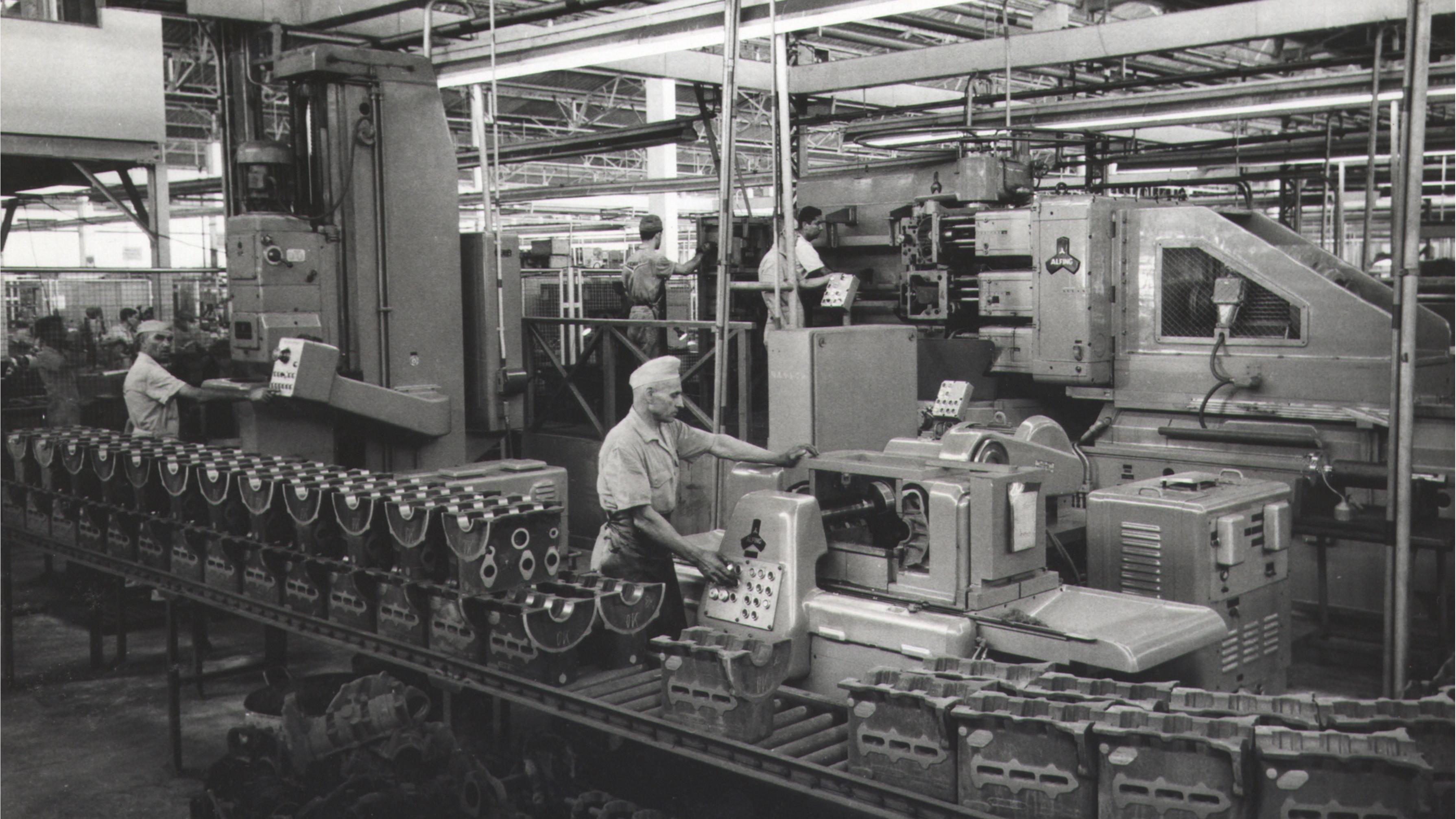 A imagem em preto e branco mostra uma linha de produ&ccedil;&atilde;o industrial dentro de uma f&aacute;brica ampla, com oper&aacute;rios trabalhando em diferentes m&aacute;quinas e equipamentos met&aacute;licos. Em primeiro plano, pe&ccedil;as mec&acirc;nicas alinhadas sobre uma esteira passam por processos de usinagem enquanto trabalhadores operam pain&eacute;is de controle e acompanham a produ&ccedil;&atilde;o. O ambiente &eacute; iluminado por estruturas industriais no teto e tem apar&ecirc;ncia de registro hist&oacute;rico de uma f&aacute;brica automotiva ou metal&uacute;rgica.