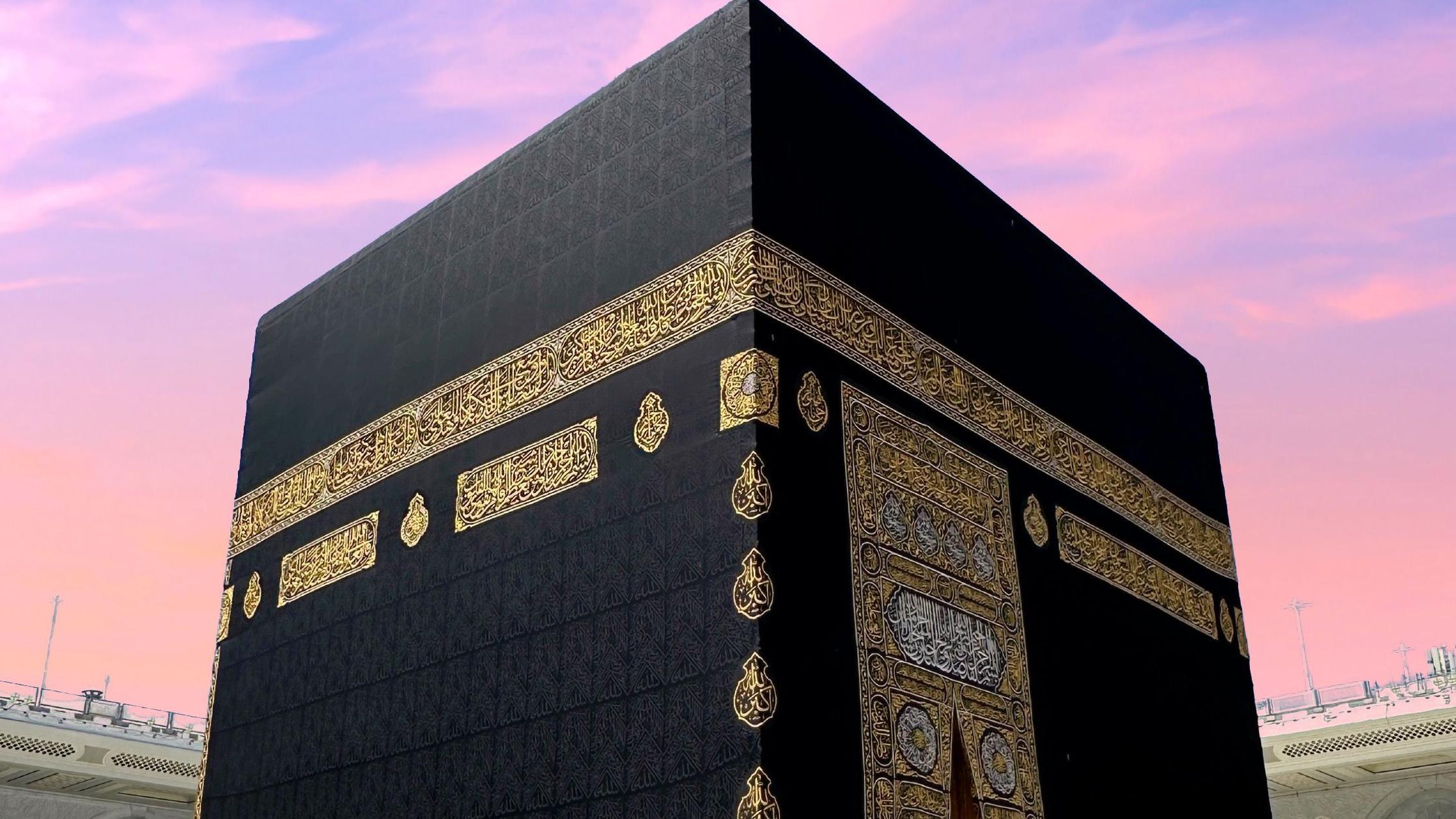 لقطة مقرّبة للكعبة، البناء المكعّب الضخم المزيّن بزخارف ذهبية وفضية، أقدس مقدسات المسلمين، داخل المسجد الحرام في مكة المكرمة، بينما تتدرّج السماء في الخلفية بين اللونين الوردي والبنفسجي.