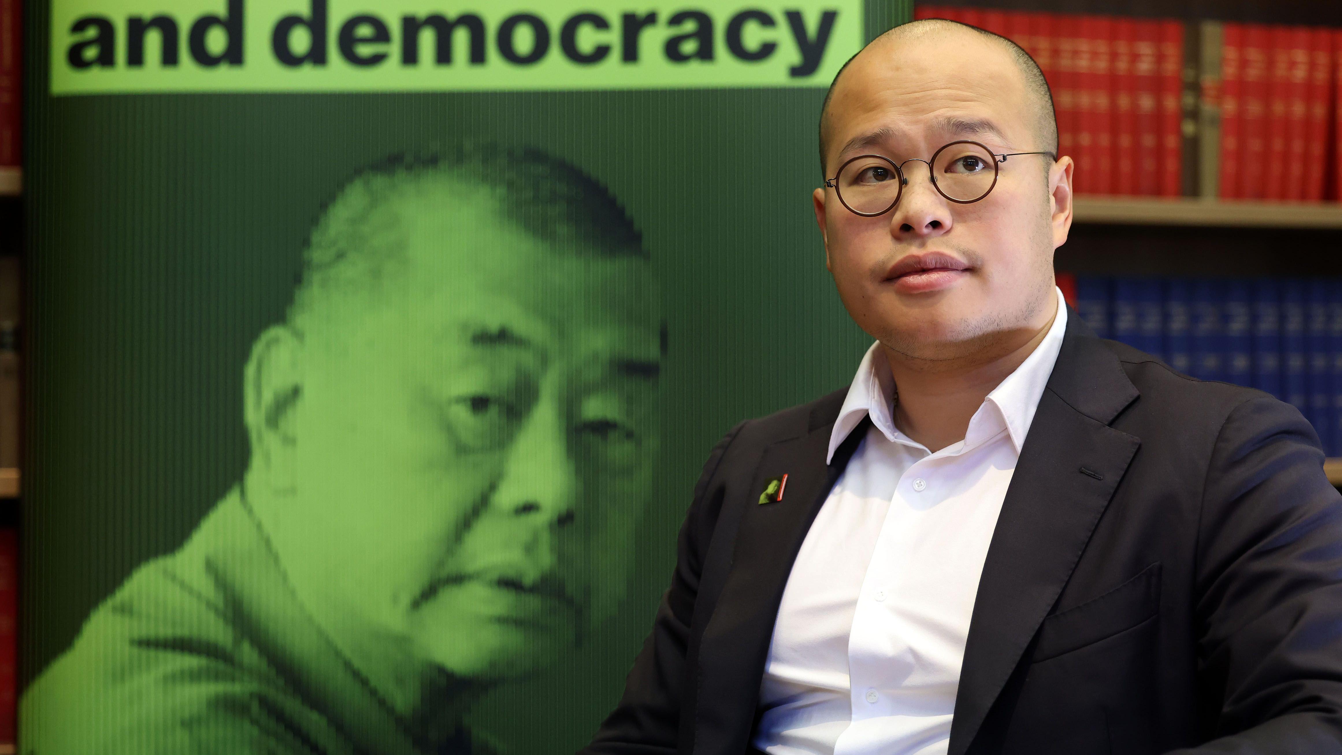 黎智英三子黎崇恩（右）在倫敦一場記者會上，坐在父親照片條幅前讓媒體記者拍照（15/12/2025）