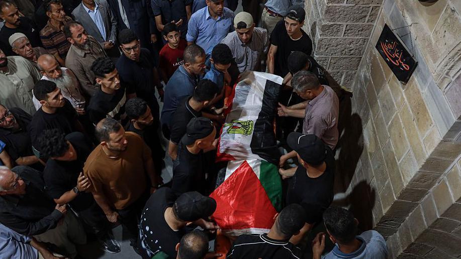 Un grupo de hombres rodean un cuerpo cubierto con la bandera palestina. 