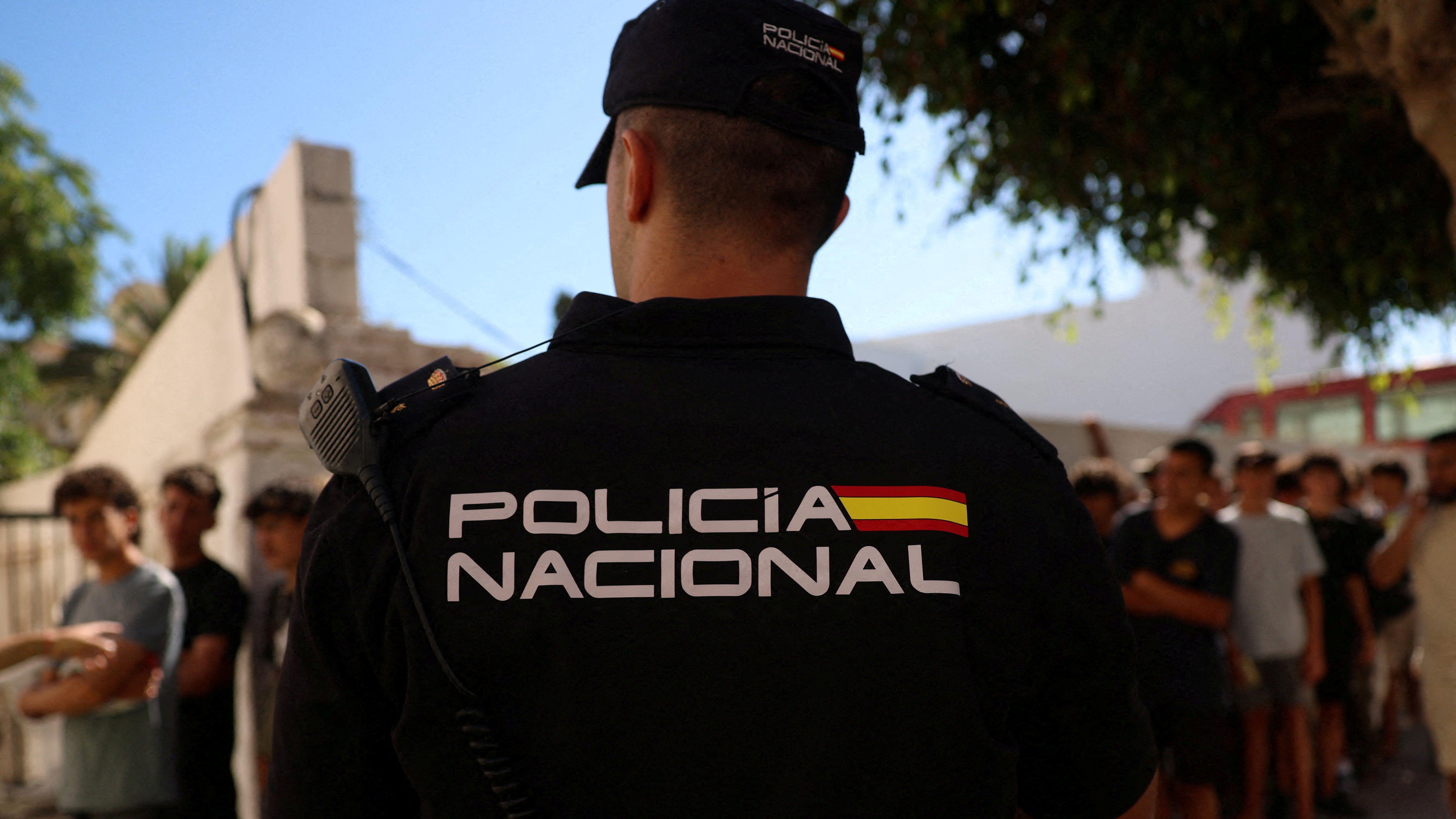 Un agente de policía monta guardia en primer plano, de espaldas a la cámara, mientras unos niños hacen cola frente a una comisaría para ser registrados ante las autoridades.