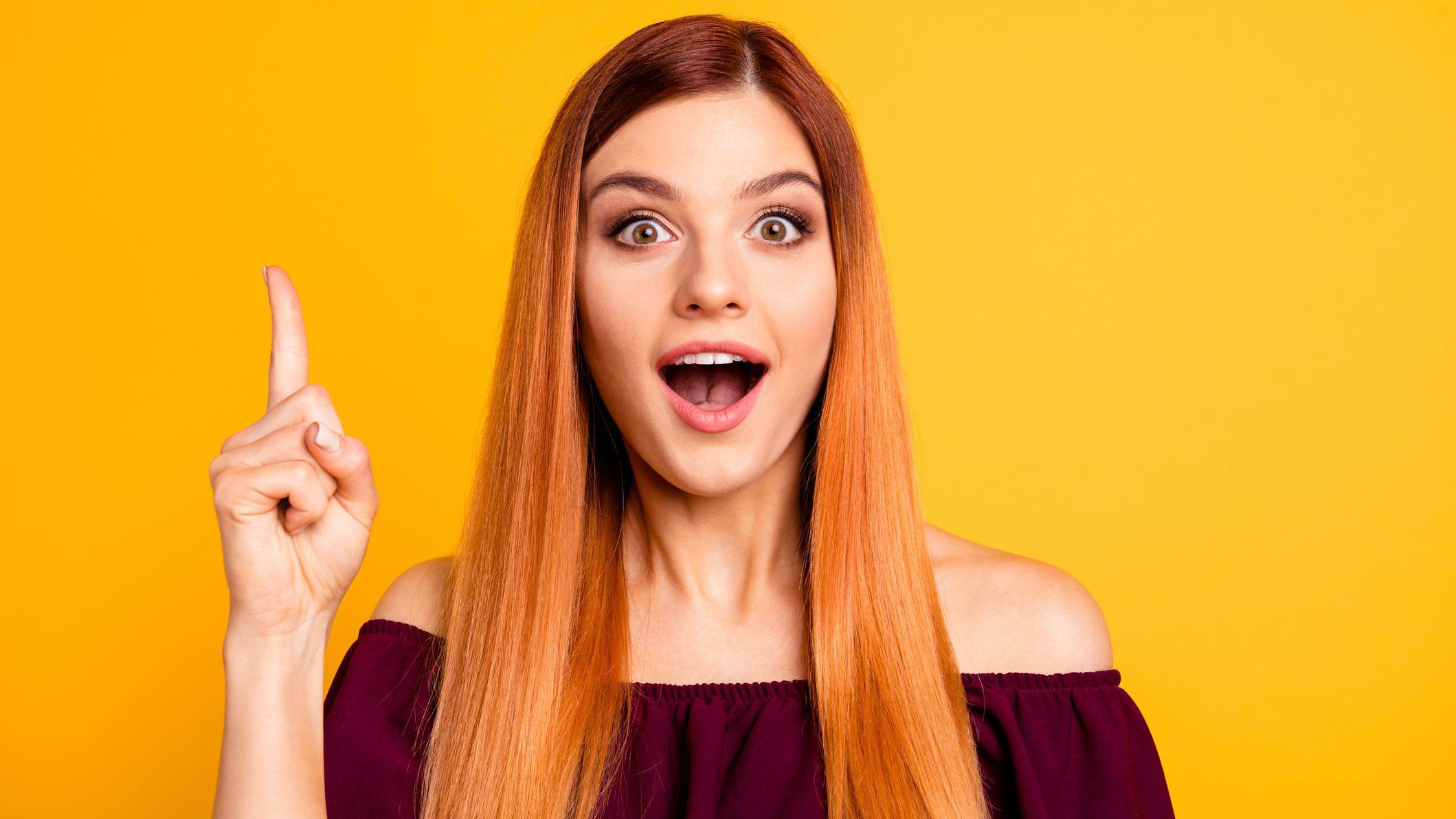 Retrato fotográfico en primer plano de una mujer joven con cabello largo rubio fresa, con una blusa vinotinto que deja ver los hombro, con la boca abierta, mirando a la cámara y sosteniendo el dedo índice levantado sobre un fondo brillante aislado.