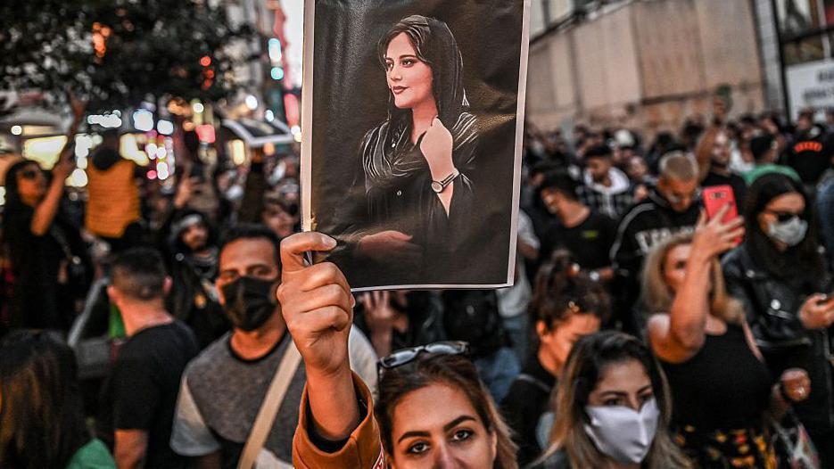 Una mujer mira a la camara y alza un retrato de la joven Mahsa Amini, en la que lleva velo. 
