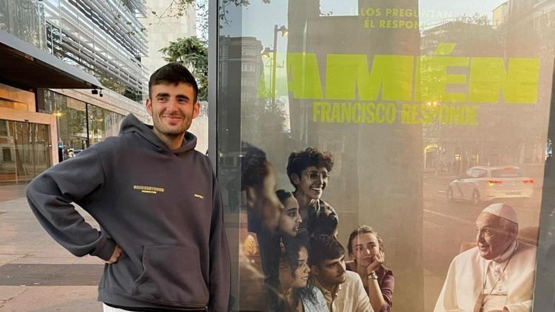 Juan Cuatrecasas Cuevas de pé, ao lado do cartaz promocional do documentário 'Amém: Perguntando ao Papa'