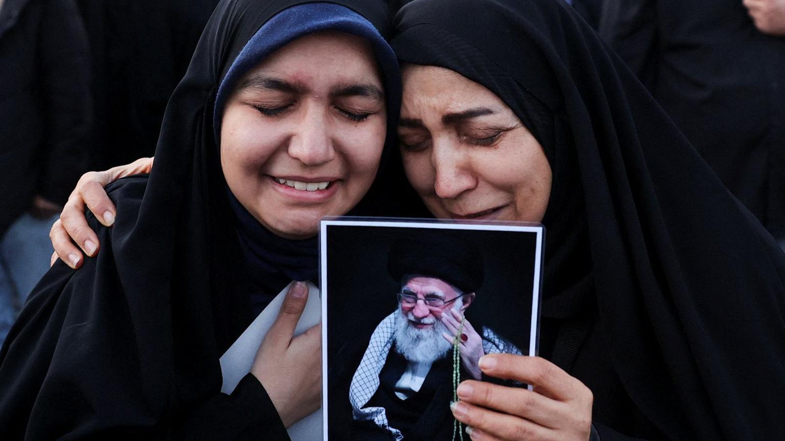 兩名女子緊緊依偎，並在伊朗最高領袖遭宣布被擊殺後舉著哈梅內伊的照片
