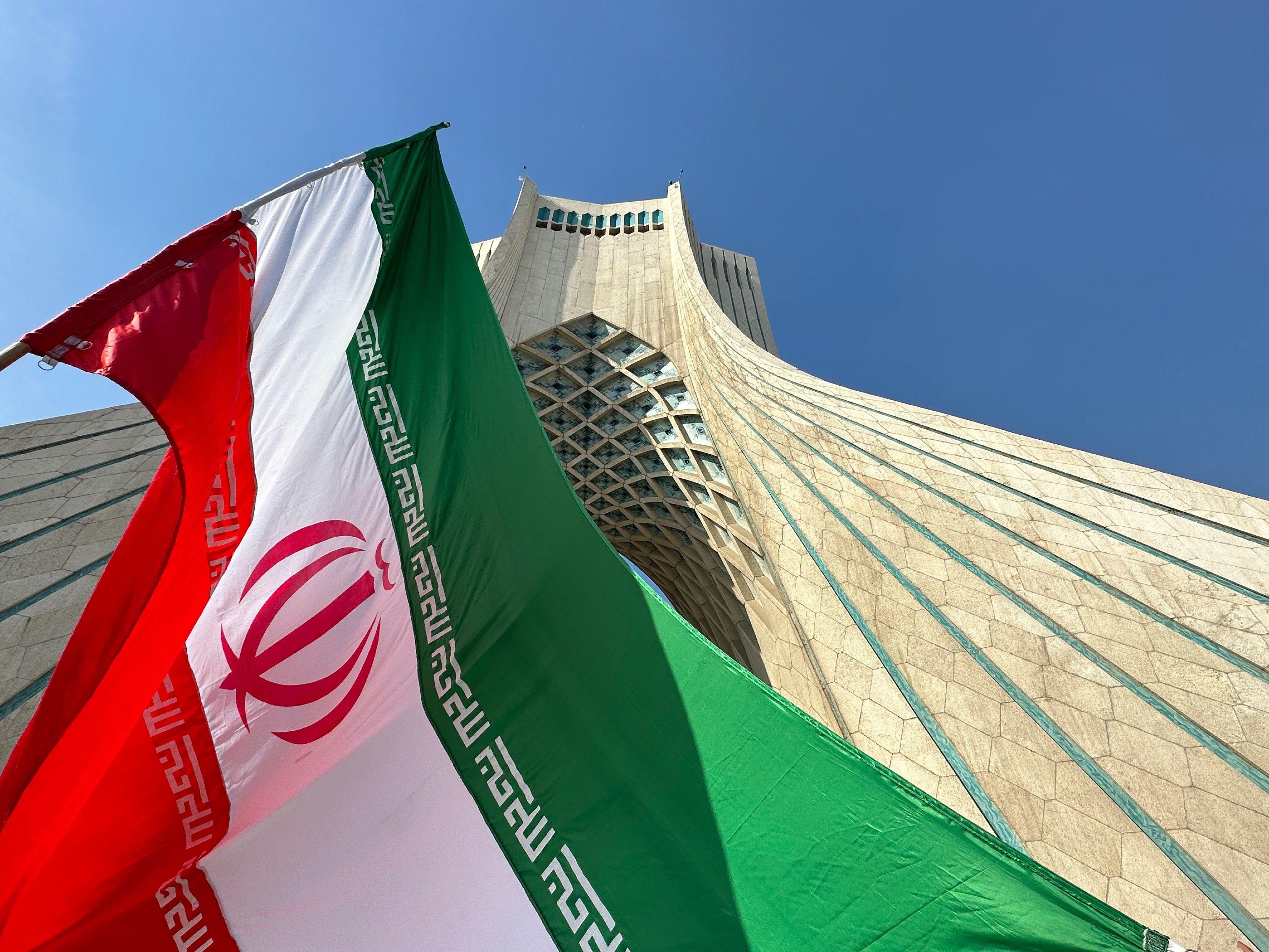 العلم الإيراني بألوانه الأخضر والأبيض والأحمر، وفي منتصفه مكتوب "الله" باللون الأحمر. وخلف العلم بناء
