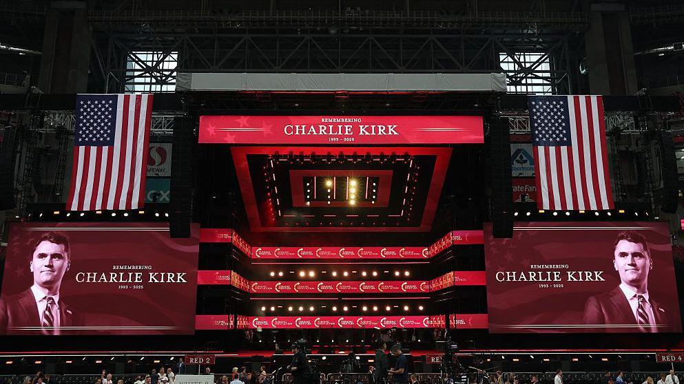 El escenario de un estadio cubierto, con grandes banderas de Estados Unidos a lado y lado y dos proyecciones de la cara y nombre de Charlie Kirk que leen: "Recordando a Charlie Kirk, 1993 - 2025"