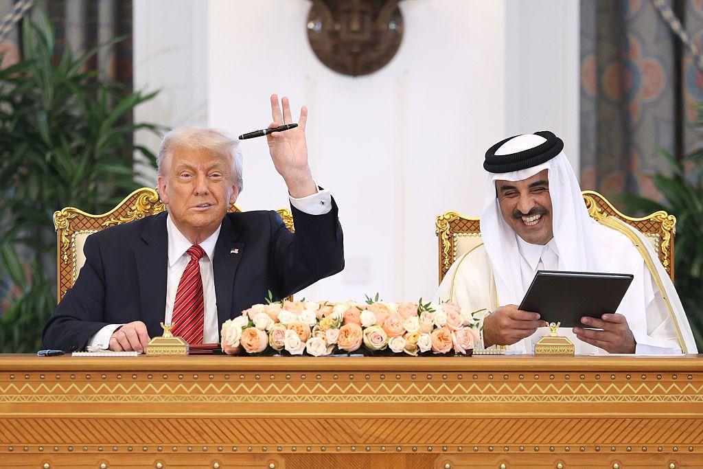 الأمير القطري مرتدياً زي بلاده الرسمي التقليدي ذي اللون الأبيض، يضحك ويمسك بأجندة سوداء اللون ويجلس بجانبه ترامب رافعاً يده التي تمسك قلماً أسود، ويرتدي بدلة رسمية سوداء وربطة عنق حمراء مخطط وقميصاً أبيض. يجلسان على طاولة ذهبية اللون مزخرفة وعليها ورود بألوان بيضاء ووردية.