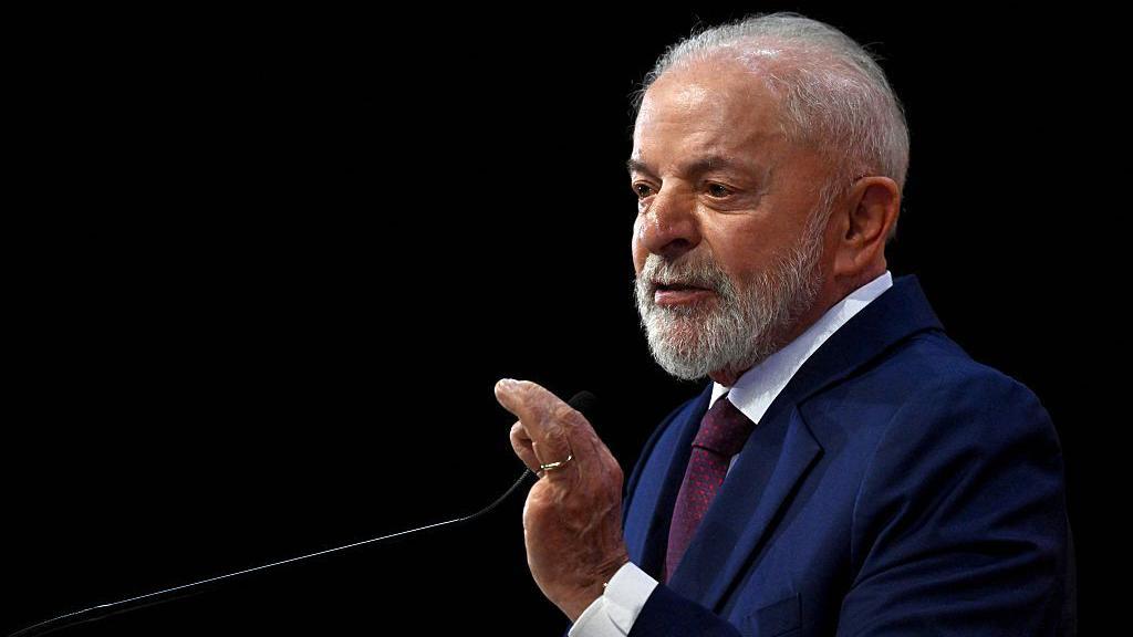 Lula &eacute; um homem branco de cabelos grisalhos. Ele est&aacute; falando ao microfone. Ele usa um terno azul.