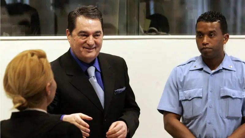 Nebojša Pavković u haškoj sudnici u crnom odelu i plavoj košulji, smeje se i pruža ruku. Pored njega je policajac u uniformi, stoji sklopljenih ruku. Ispred Pavkovića je žena, u crnoj garderobi, okrenuta leđima, sa narandžastom kosom