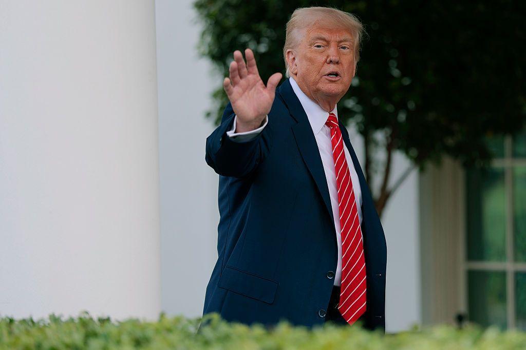 يلوّح الرئيس الأمريكي دونالد ترامب بيده من البيت الأبيض. وهو يرتدي بدلة كحلية وربطة عنق حمراء مخططة باللون الأبيض.