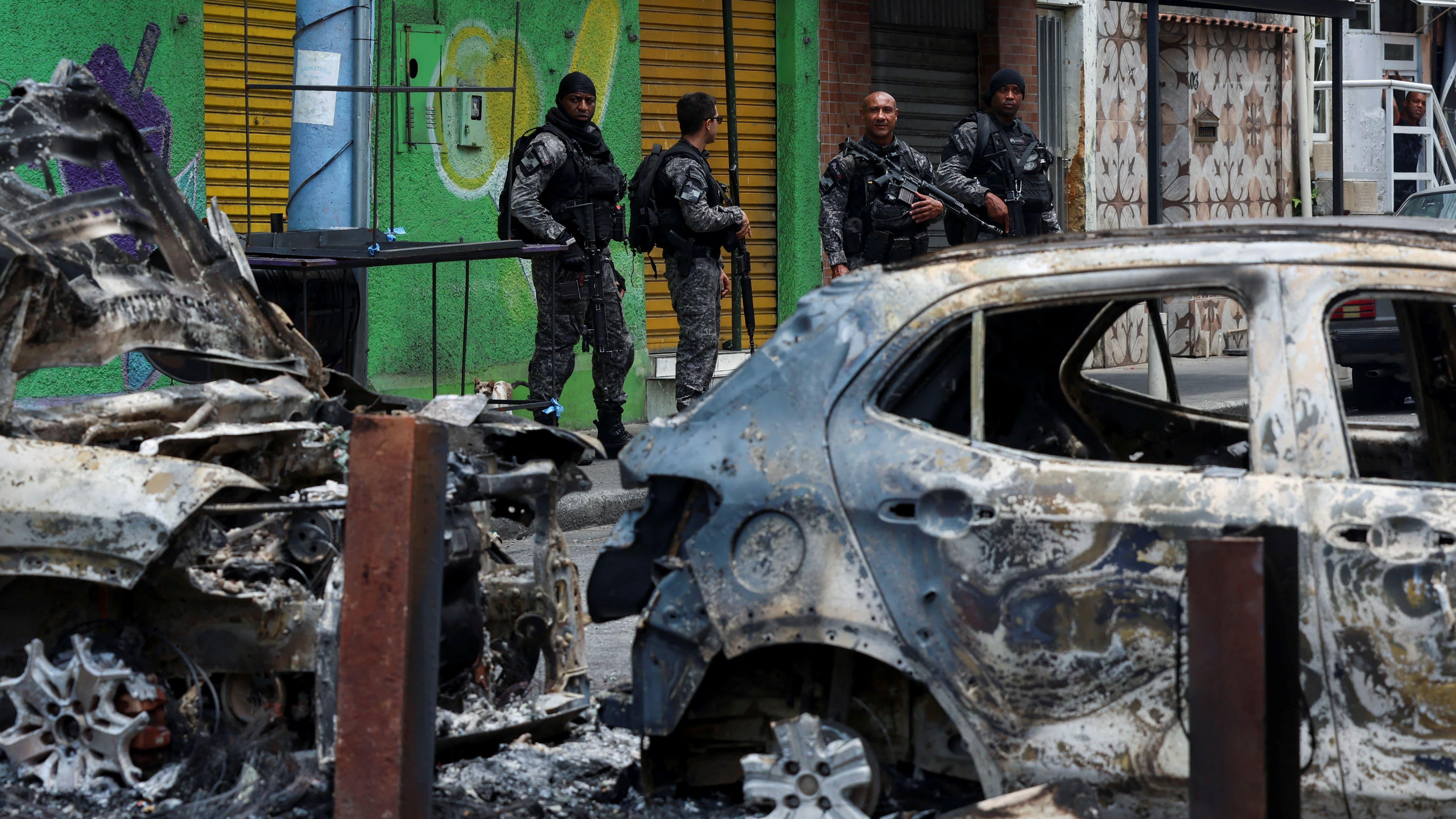 Policias fortemente armados observam carca&ccedil;as de carros encendiados