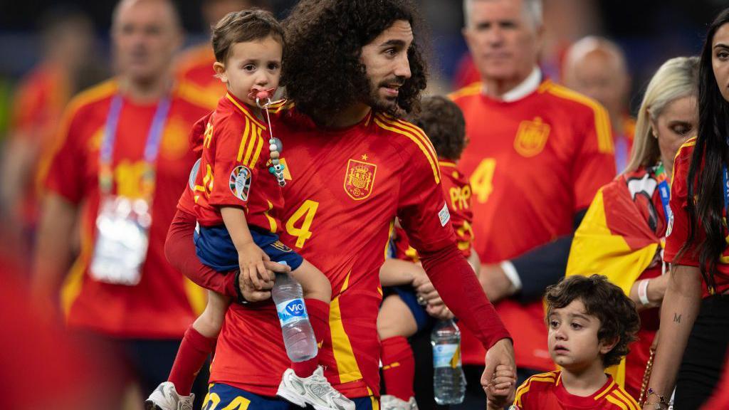 Cucurella con la camiseta roja de España sujeta con una mano a su hijo pequeño y con otra a Mateo