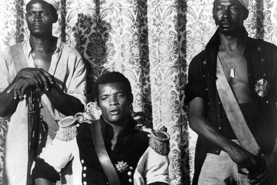 Una escena de la película "Queimada". Evaristo Márquez, como José Dolores, está sentado con su uniforme de charreteras. Atrás hay dos hombres con armas, que interpretan esclavos rebeldes