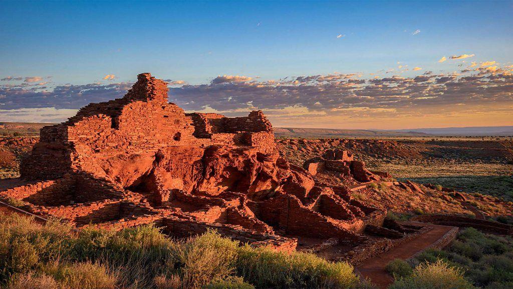 Ruinas de una estructura de piedra rojiza en un paisaje desértico, iluminadas por la luz del atardecer bajo un cielo parcialmente nublado.