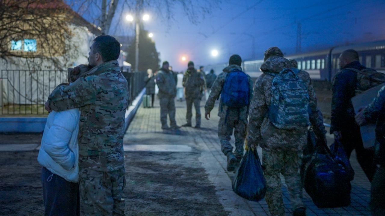 Un hombre con uniforme militar abraza a una mujer en una estación de tren. A su lado, pasan otros militares cargados con mochilas y otros bultos.